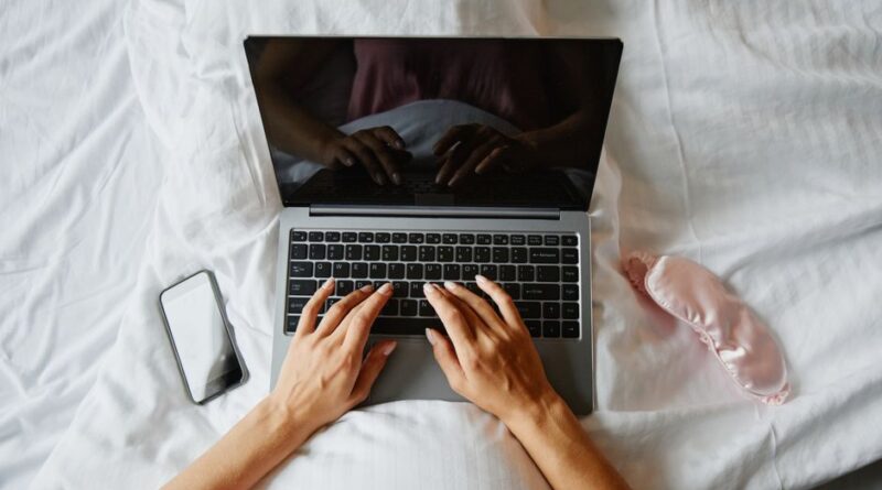 Person lying in bed using a laptop, with a phone and sleep mask nearby, creating a cozy indoor work environment. - Photo by Marcus Aurelius on Pexels